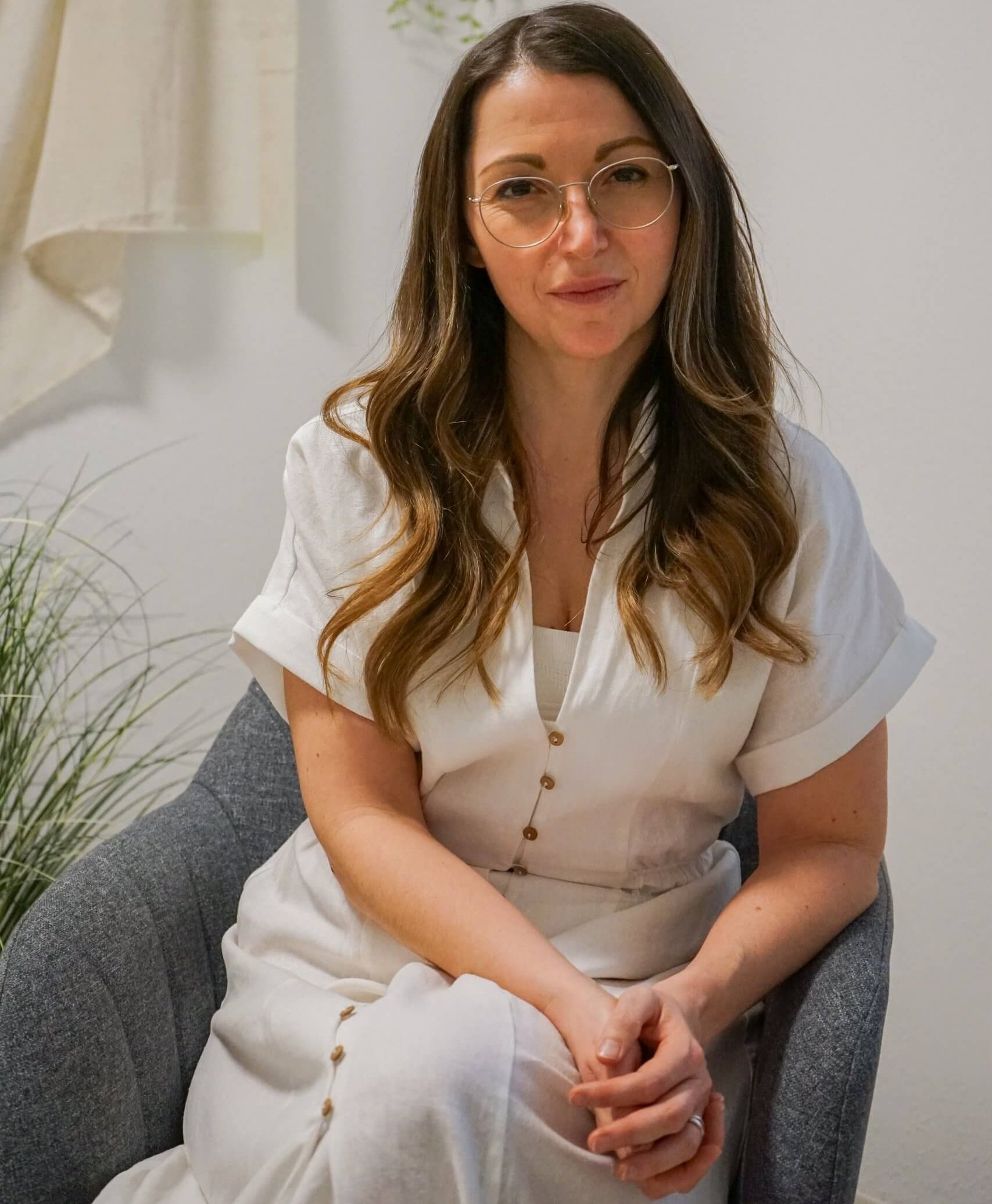 Eine Frau mit langen braunen Haaren und Brille sitzt auf einem grauen Stuhl, trägt ein weißes Kleid, im Hintergrund Pflanzen und Stoff